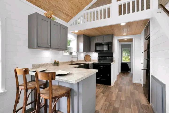 Modern kitchen with vaulted wooden ceiling, gray cabinets, black appliances, and stone countertops. Wooden chairs at a breakfast bar add warmth. Cozy and inviting.