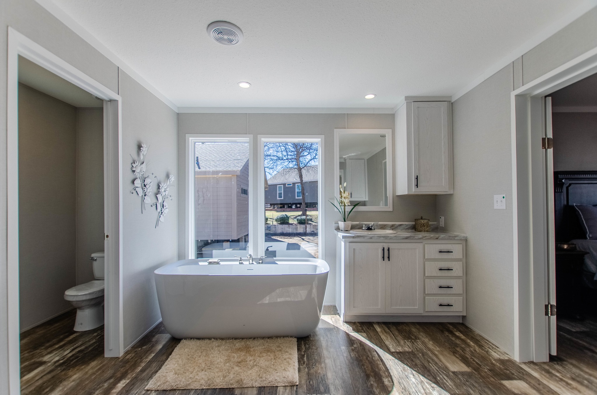Modern bathroom with a standalone bathtub under large windows, offering a view outside. Features sleek cabinetry and dark wood flooring, creating a serene atmosphere.