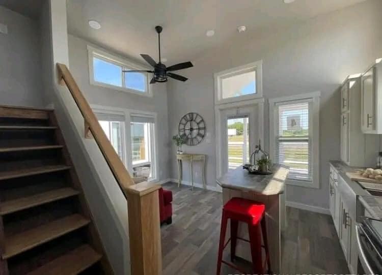 Bright kitchen and living area featuring large windows, wood flooring, and a stairway. A red stool sits by a kitchen island, and a large wall clock adds charm.