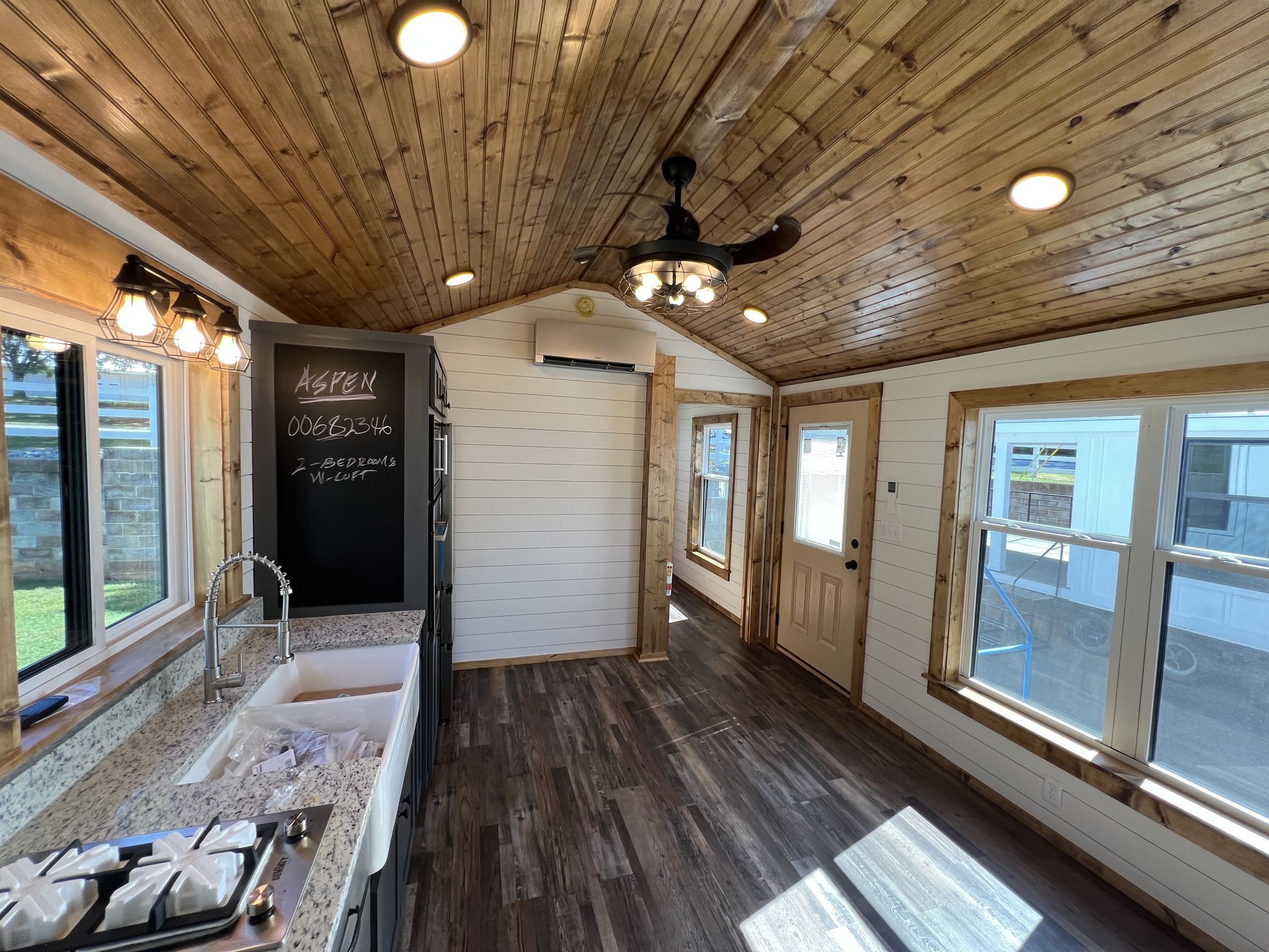 Cozy tiny home interior with rustic wooden ceiling and floors, modern lighting, and large windows. A chalkboard lists property details. Warm and inviting ambiance.