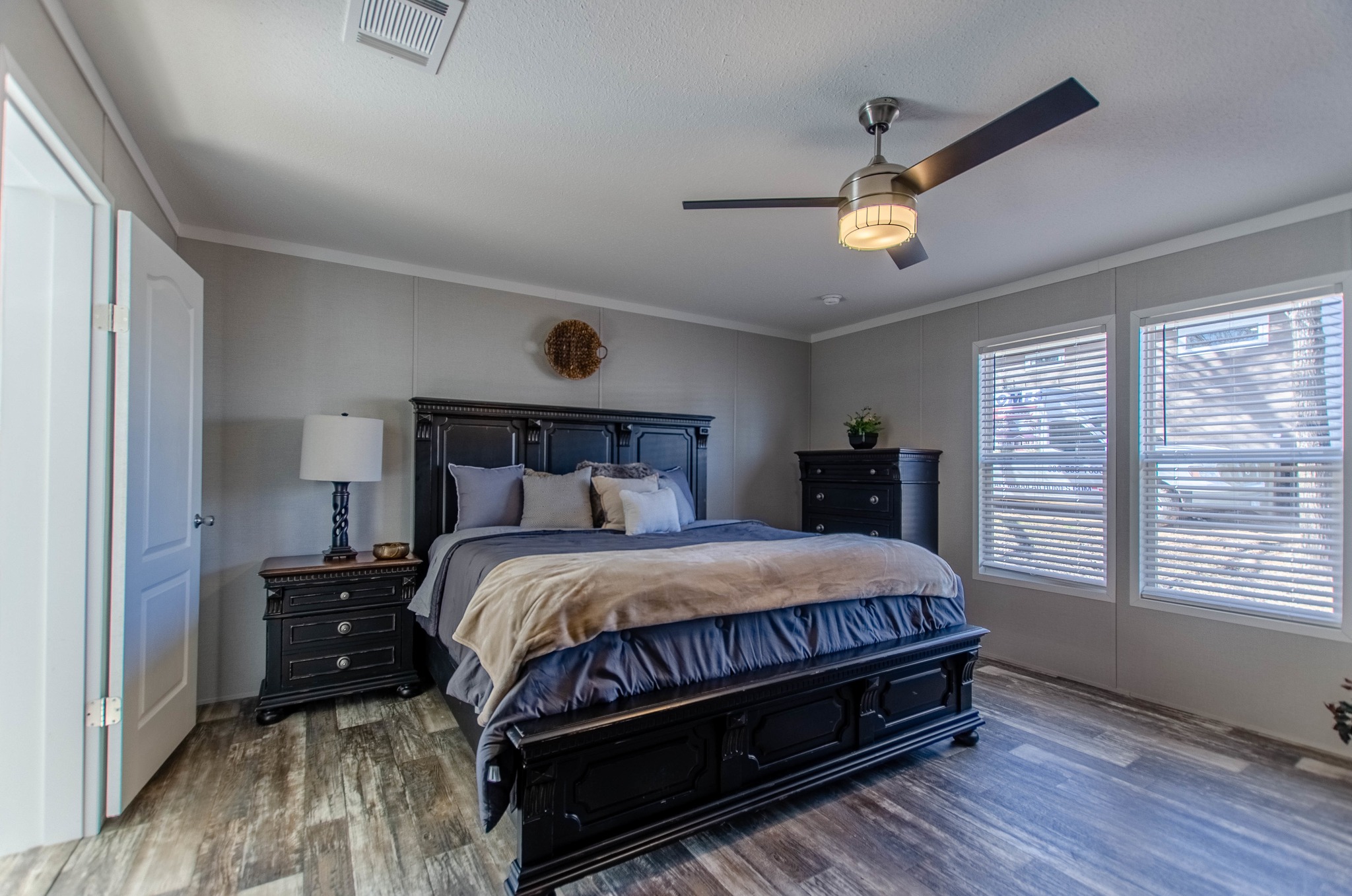 A cozy bedroom with a dark wood king-sized bed, matching nightstands, and a dresser. Light gray walls, large windows, and ceiling fan add brightness.