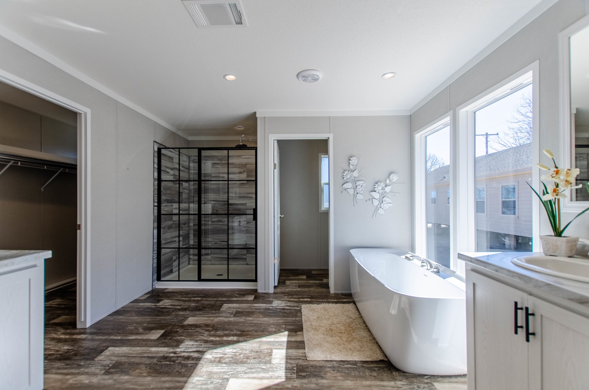 Spacious bathroom with large glass shower, freestanding white tub, and sunlight streaming through three windows. Modern decor with floral wall art.