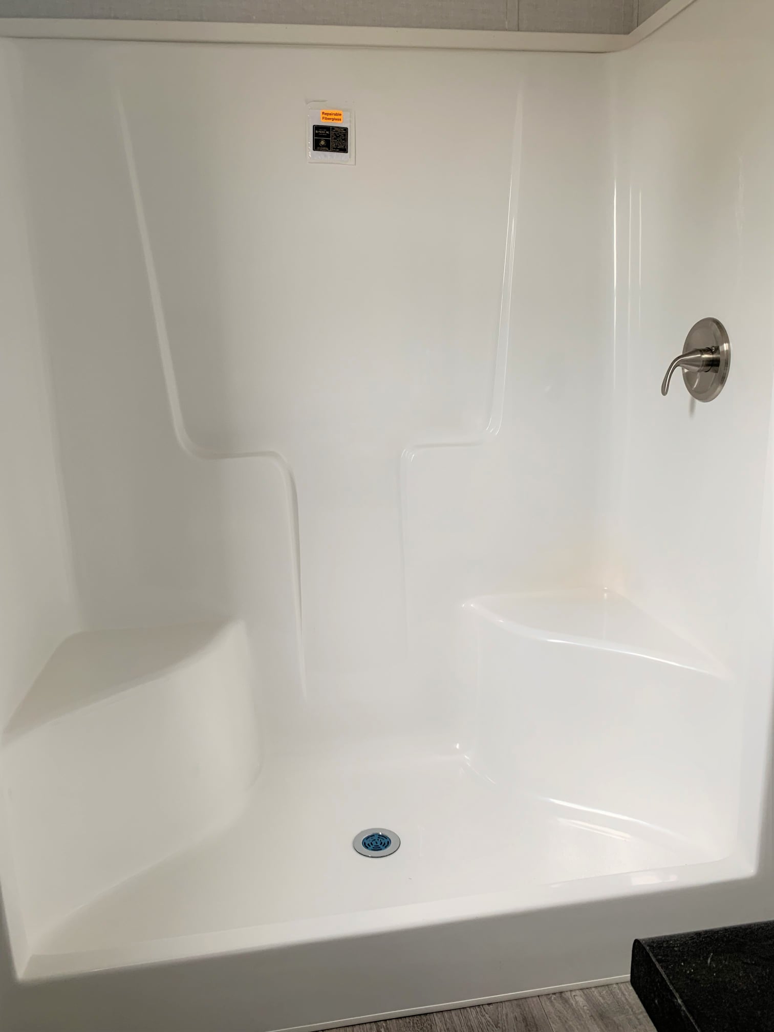 A white shower cubicle with a smooth finish features a silver faucet on the right wall and built-in corner seating. The floor has a centered drain.