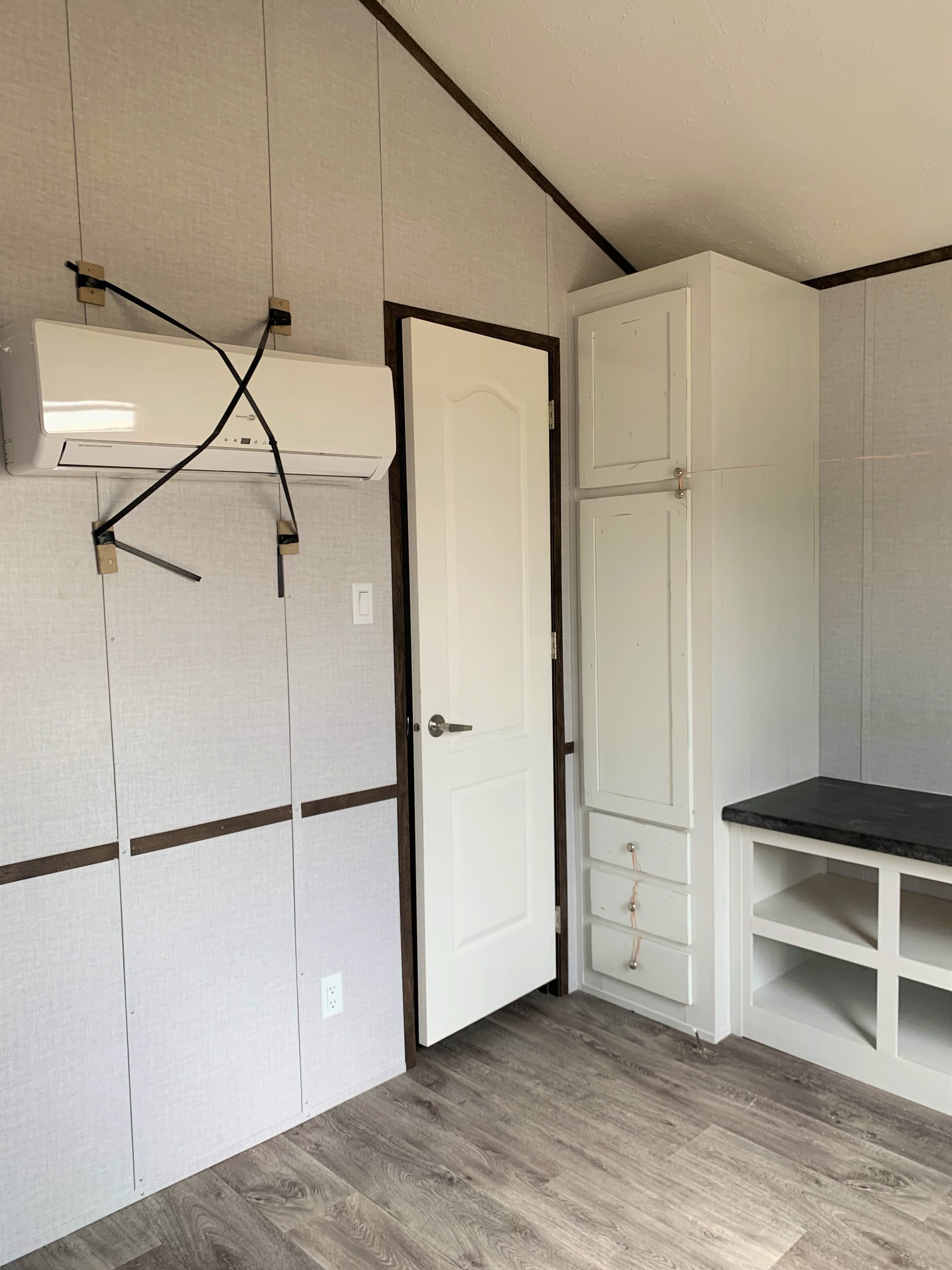 Small, empty room with light gray walls, a white door, and wood flooring. Wall-mounted air conditioner with straps, white cabinets, and shelves visible.