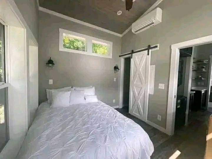 Cozy bedroom with a white, plush bed next to windows. A barn-style sliding door leads to an adjoining room. Soft lighting and neutral walls create a serene ambiance.