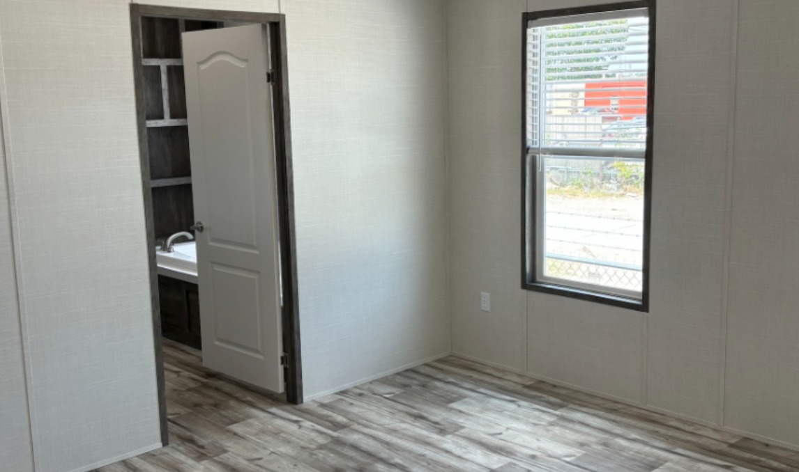 Simple, unfurnished room with light-colored walls, wood floor, window with blinds, and an open door leading to a bathroom with visible sink.