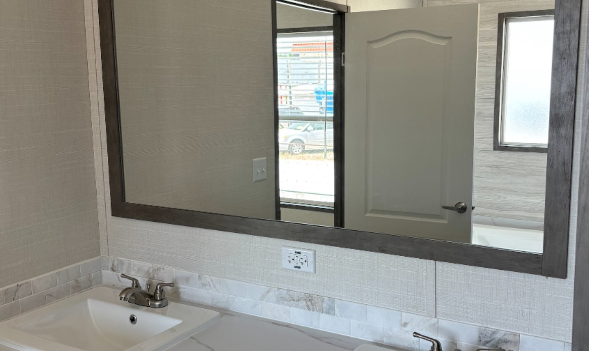 A modern bathroom with a large mirror reflecting a partially open door. The sink has a metallic faucet, set against a white marble countertop and light-colored walls.