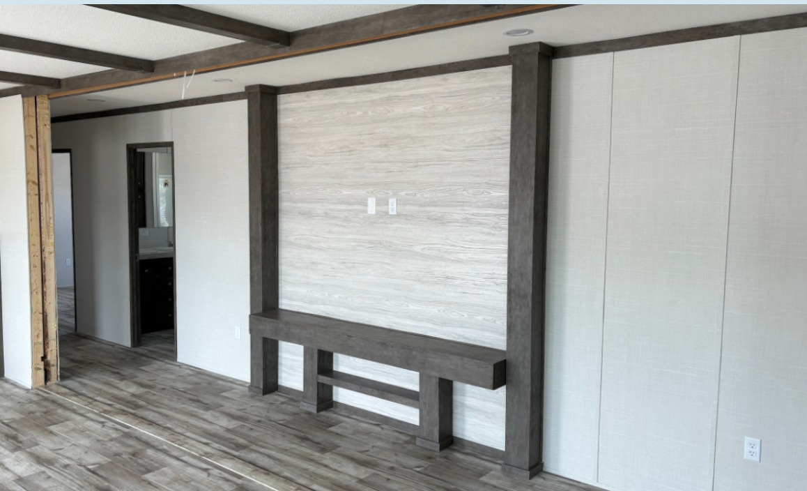 A modern, empty room features light wood flooring, white walls with dark wood trim, and a built-in wooden accent panel. A doorway leads to another room.