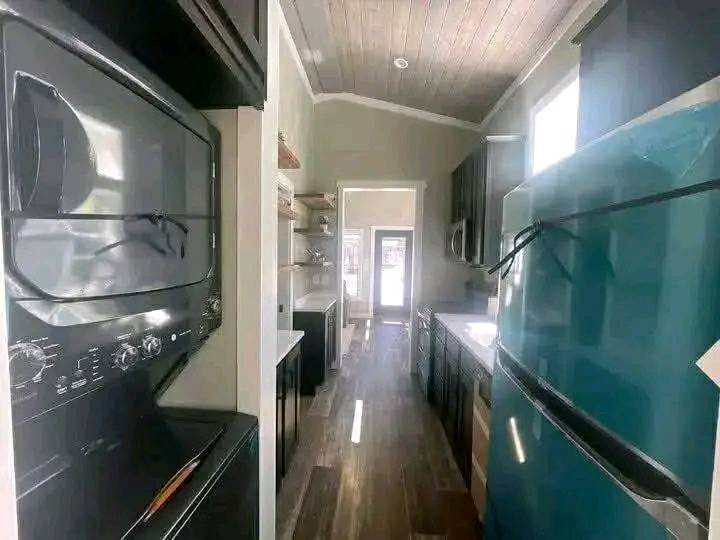 Narrow kitchen with wood ceiling and floor, featuring a teal refrigerator, dark cabinets, and a stacked washer-dryer unit, creating a cozy vibe.
