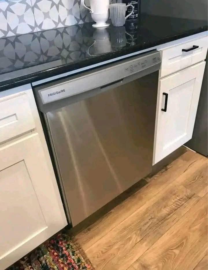 Stainless steel Frigidaire dishwasher in a modern kitchen, flanked by white cabinets with black handles, black countertop, and geometric backsplash.