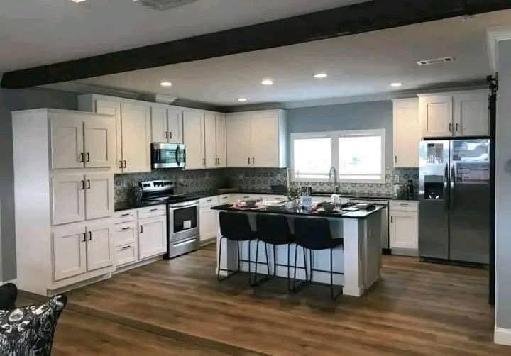 Modern kitchen with white cabinets, stainless steel appliances, and a central island with three black stools. Wooden floor and patterned backsplash.