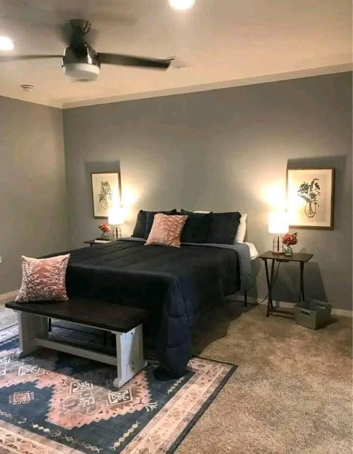 A cozy bedroom with a dark bedspread, pink pillows, and side tables with lit lamps. Gray walls, framed art, ceiling fan, and patterned rug create a serene atmosphere.