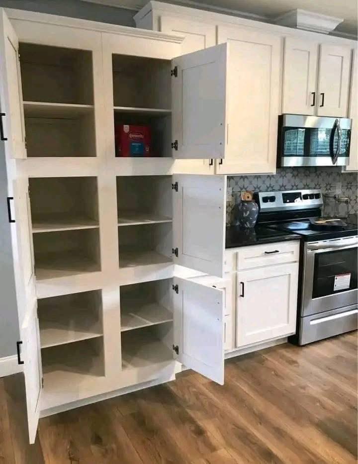 Open white kitchen pantry with multiple empty shelves, one box of cereal on the top shelf. Next to stainless steel stove and microwave, wood floor.