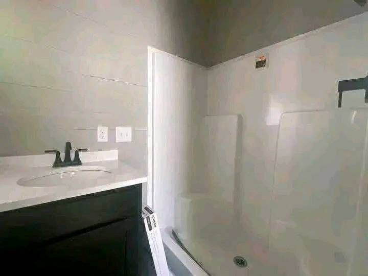 A modern bathroom features a dark vanity with a sleek black faucet and a white countertop. Adjacent is a spacious white shower, offering a clean, minimalist feel.