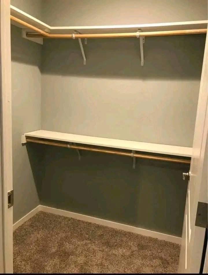 Empty walk-in closet with two wooden garment rods and a shelf against gray walls. Soft carpet covers the floor, creating a simple and clean look.