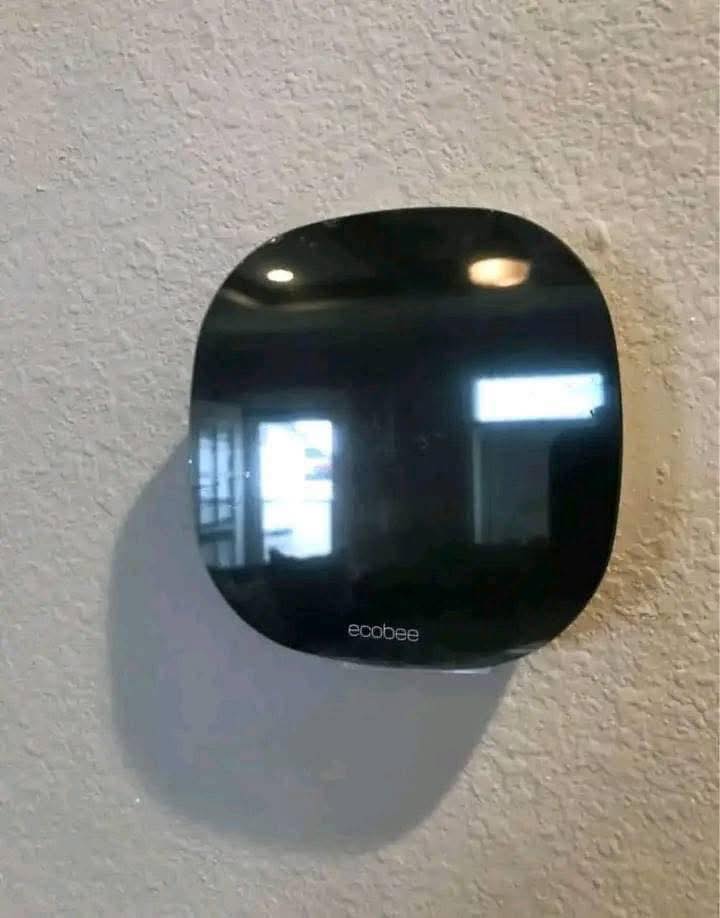 A black Ecobee smart thermostat mounted on a light, textured wall. The screen is dark, reflecting light from windows and ceiling lights in the background.