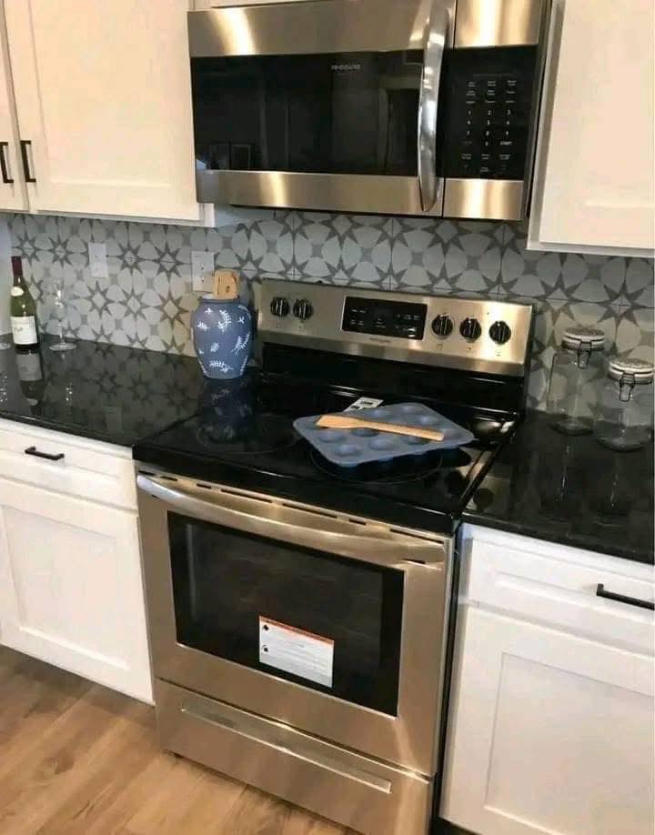 A modern kitchen features a stainless steel oven and microwave. Black countertops, geometric backsplash, and a blue vase add sleek touches.