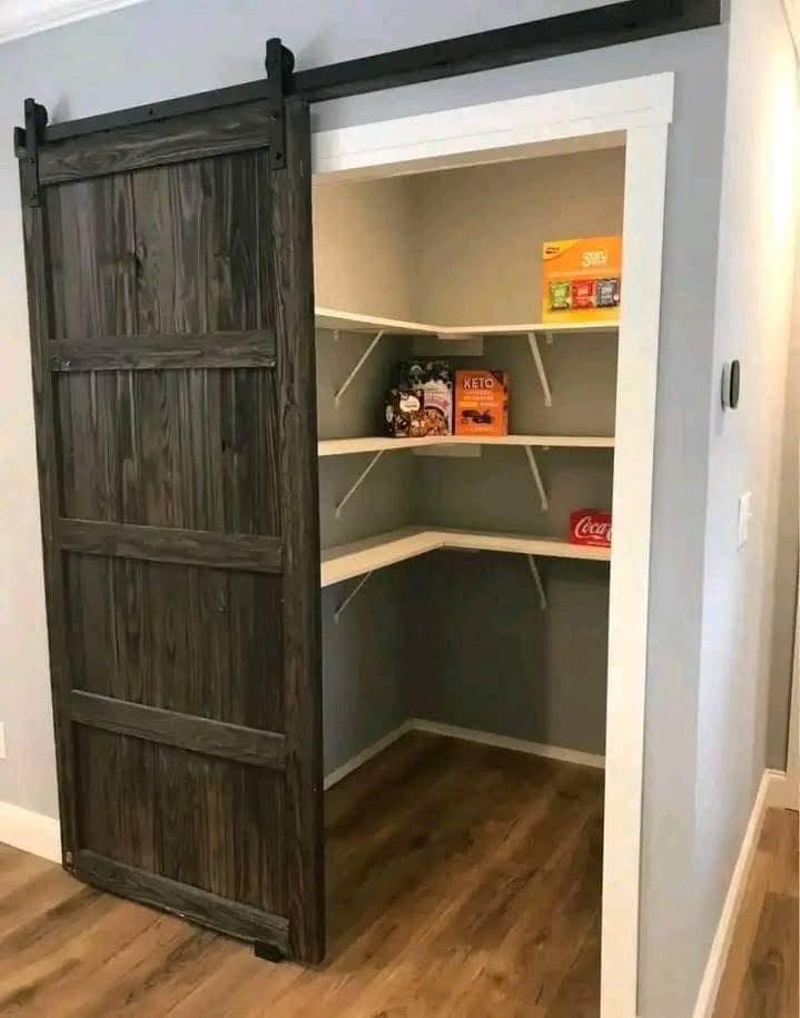 A small pantry with wooden shelves holds snack boxes and a soda bottle. A rustic sliding barn door reveals the neatly organized space.