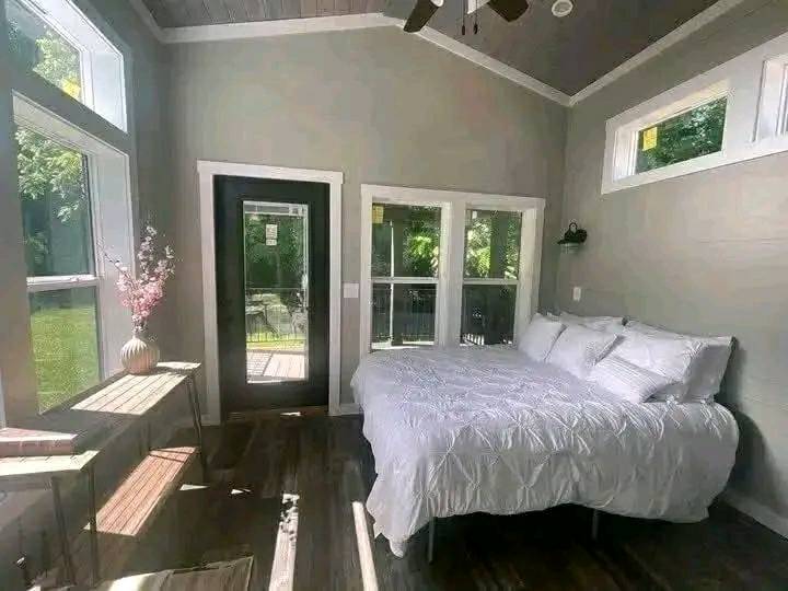 A cozy bedroom with gray walls and large windows showcasing a lush outdoor view. It features a bed with white linens, bright natural light, and a minimalist vibe.