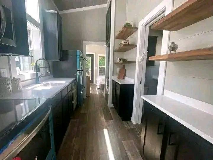 Narrow kitchen with dark cabinets, white countertops, and open wooden shelves. Sunlight streams in through a large window, creating a cozy ambiance.