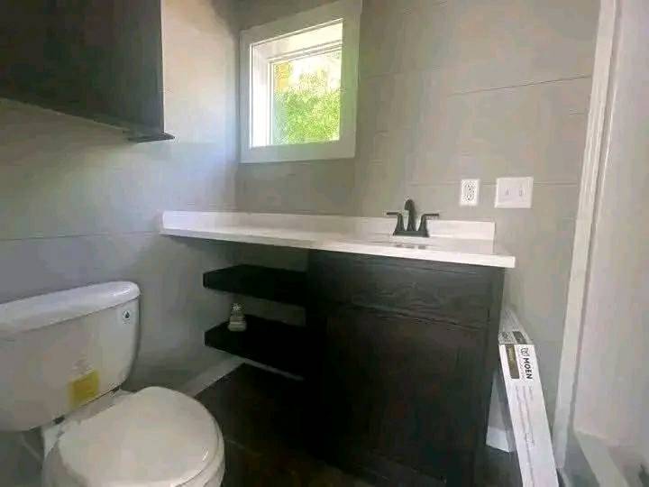 A compact bathroom with a dark wood vanity and white countertop under a small window. The toilet is adjacent, with a dark faucet and shelves nearby. Bright and serene ambiance.
