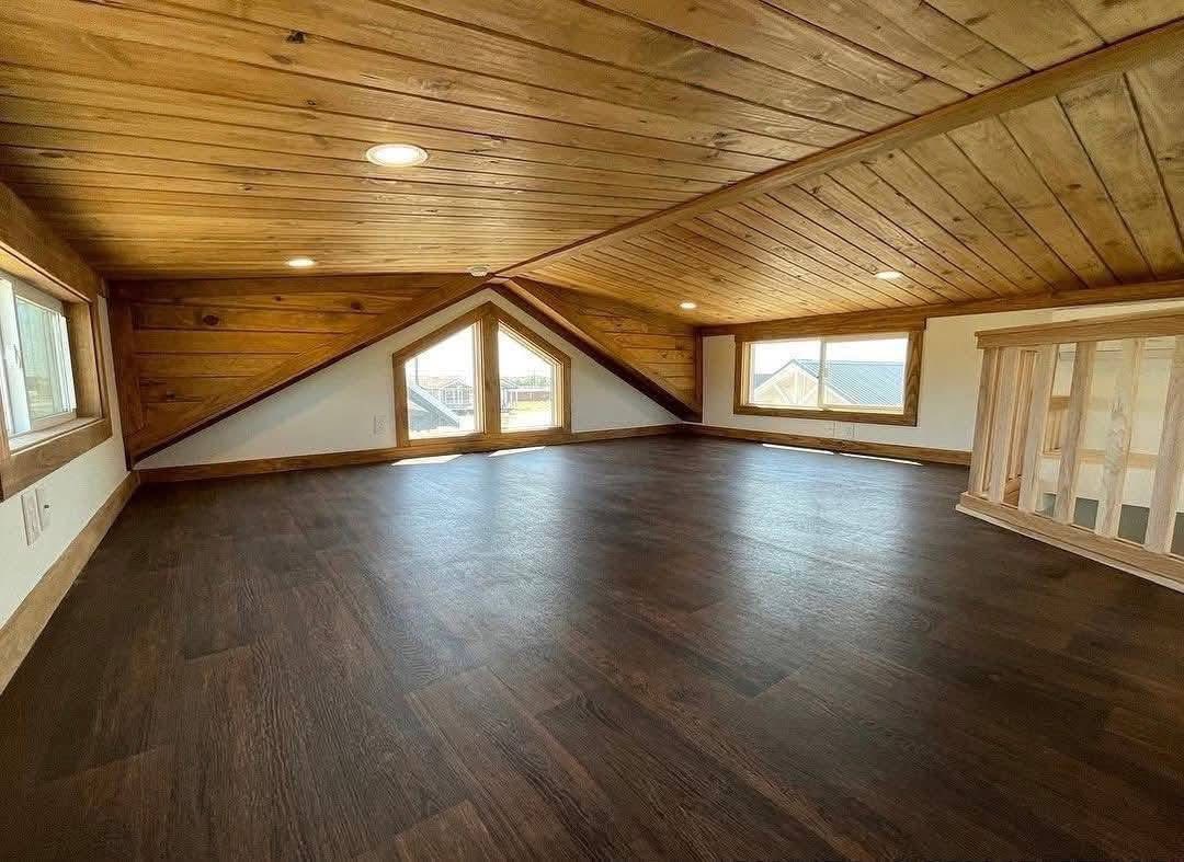 Spacious attic room with sloped wooden ceiling and dark hardwood floor, featuring three triangular windows and a railing on the right. Bright, cozy ambiance.