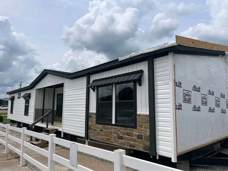 Modern modular home partially wrapped, features white siding and stone accents. Overcast sky and a white fence create a calm, residential setting.