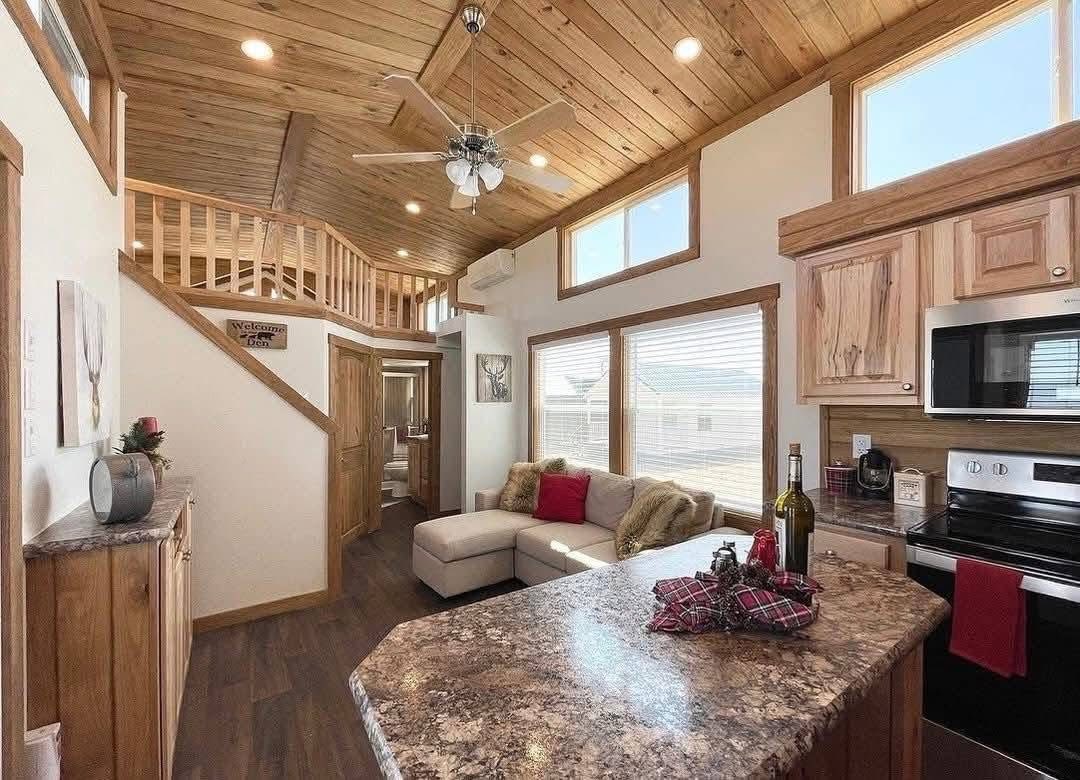 Cozy cabin interior with a wooden ceiling, modern kitchen, and a beige sectional sofa with red cushions. A loft, ceiling fan, and sunlight enhance warmth.