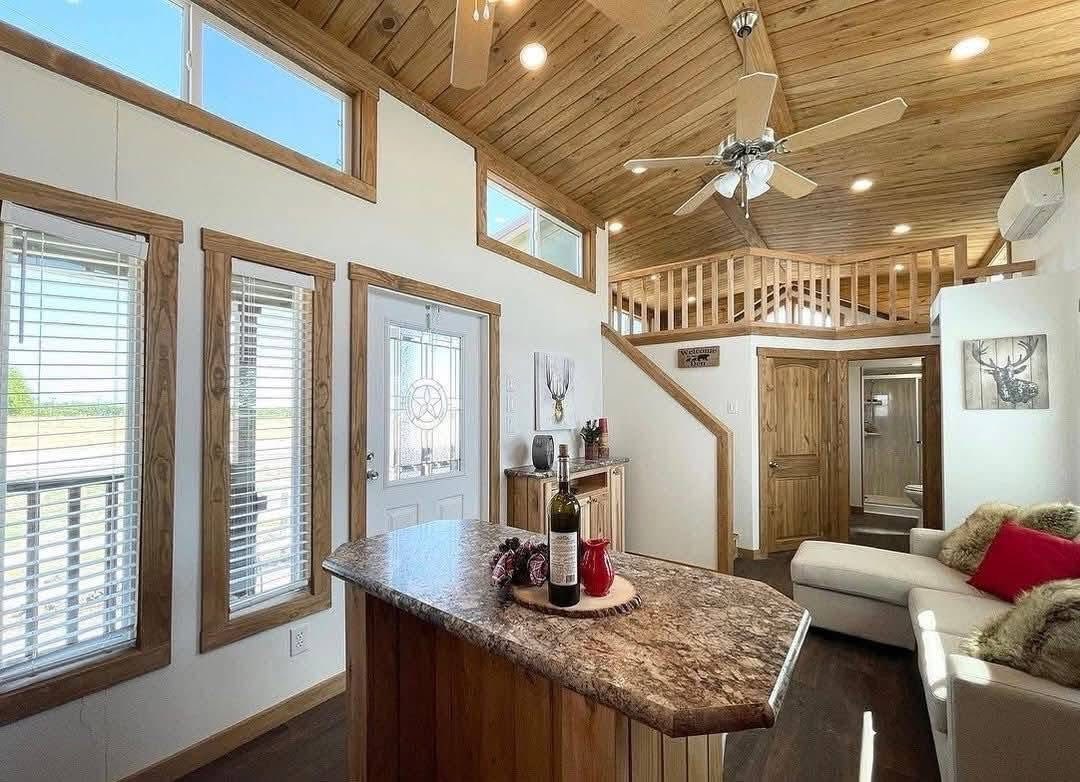 Cozy cabin interior with a vaulted wooden ceiling, ceiling fan, and natural light. A kitchen island with wine and decor, white couch with red pillows.