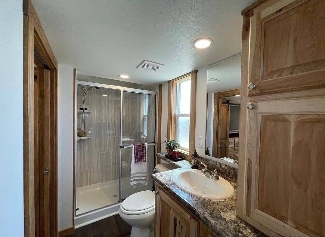 Compact bathroom with wooden cabinets, featuring a glass-enclosed shower, vanity with sink, toilet, and small plant by the window. Bright and cozy.