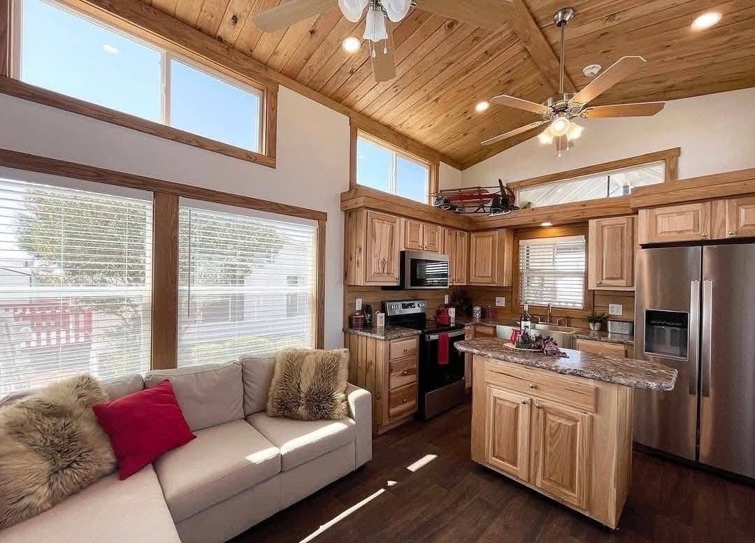 Cozy living space with wooden ceiling and natural light. Features a beige couch with red and fur cushions, modern kitchen, and ceiling fan. Warm and inviting.