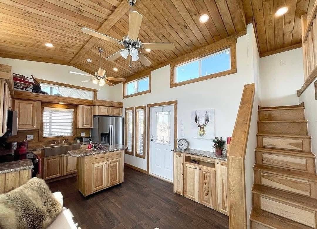 Warm, rustic tiny home interior with wood paneling and high ceilings. Features a small kitchen, cozy loft, and staircase. Natural light fills the space.