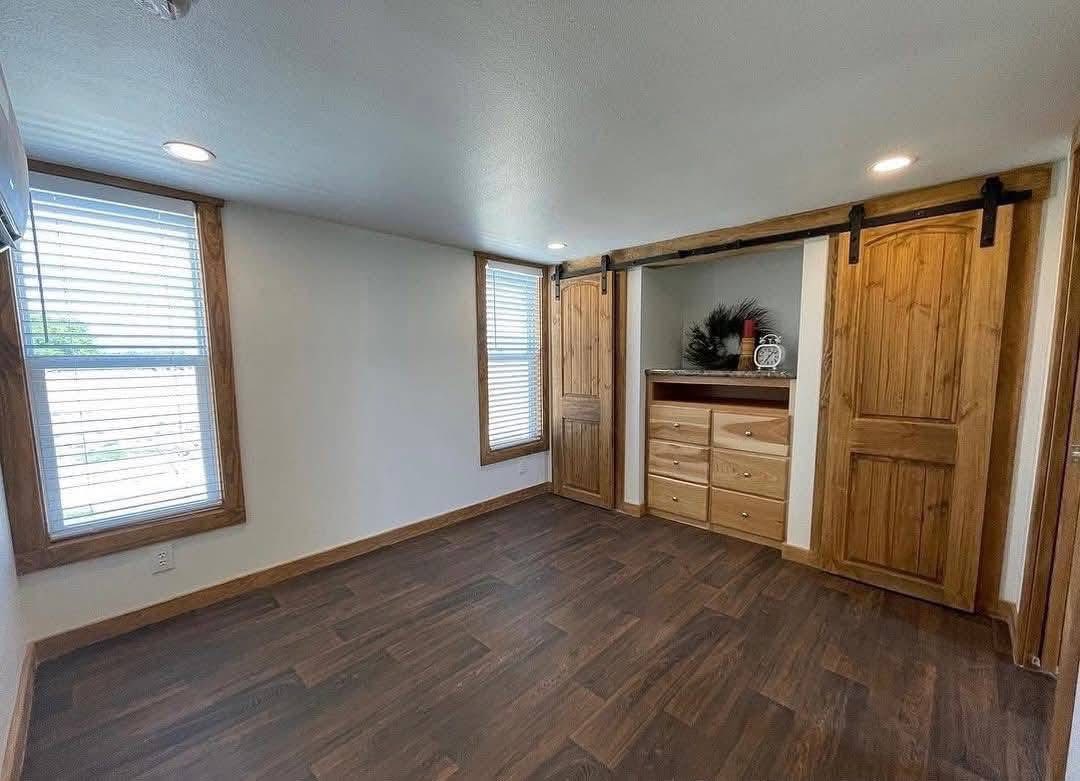 Empty room with dark wood flooring, white walls, and large windows with wooden trim. Features sliding barn doors open to shelves and drawers. Bright, cozy atmosphere.