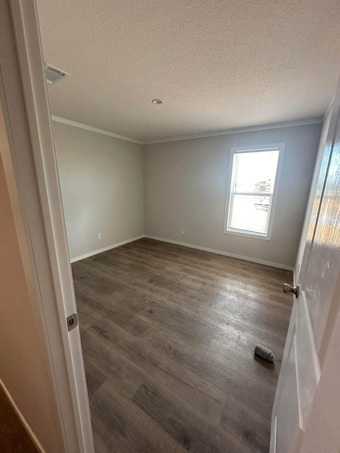 Empty room with light gray walls, a single window allowing natural light, and dark wood laminate flooring. A sock is on the floor, adding a casual tone.