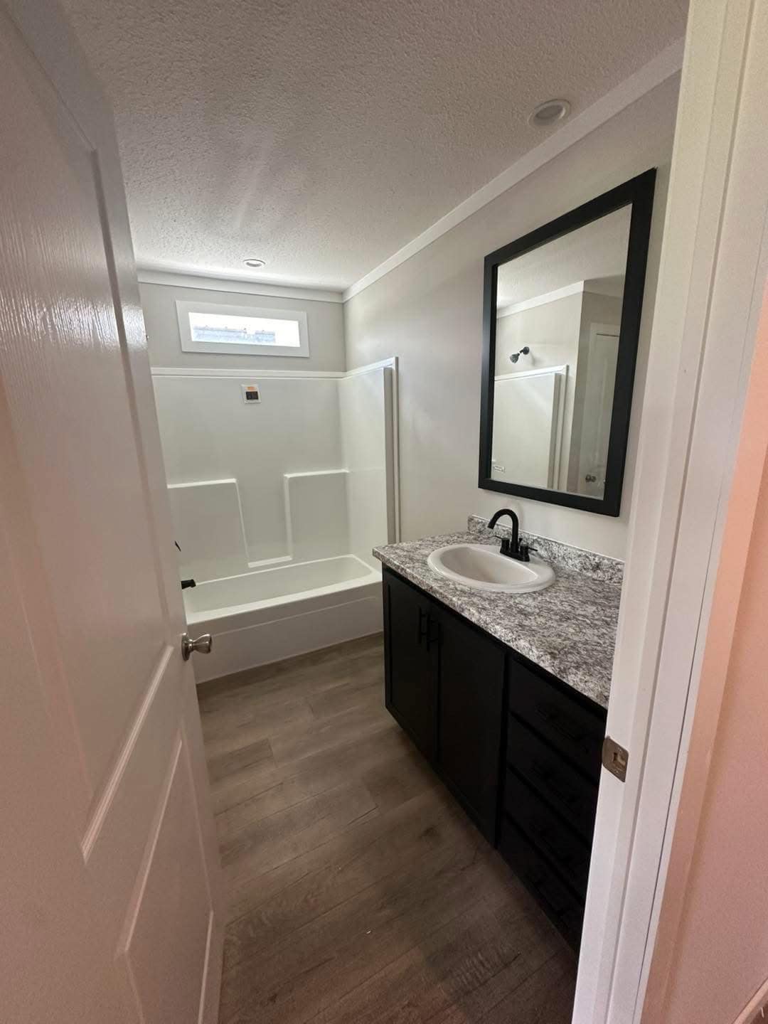 Compact bathroom with a white bathtub and shower, gray marble vanity, black-framed mirror, and wood floors. Bright and clean atmosphere.