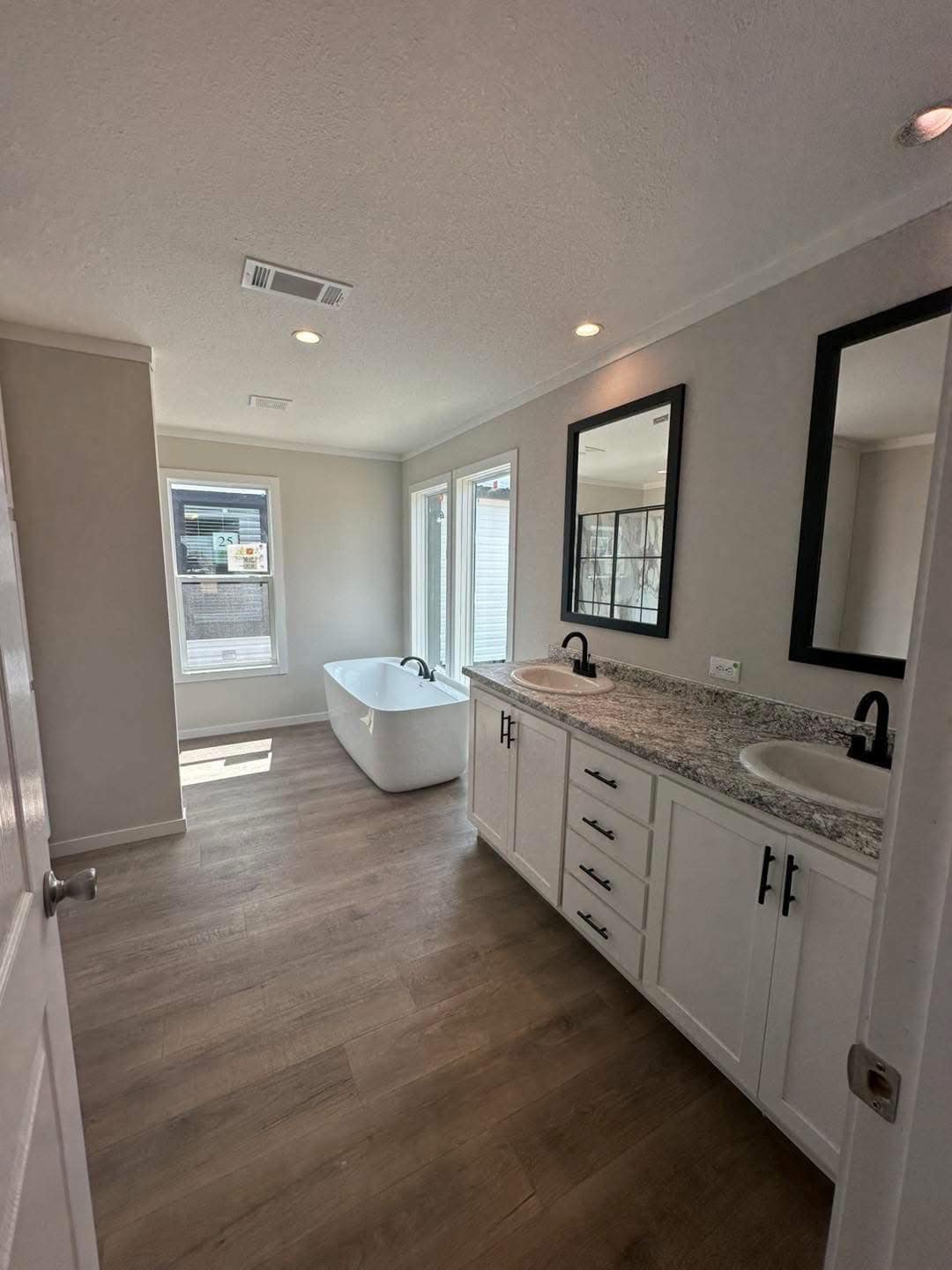 Bright bathroom with wooden floor, featuring a white freestanding tub by large windows. A marble double sink vanity has black-framed mirrors above, creating an elegant vibe.