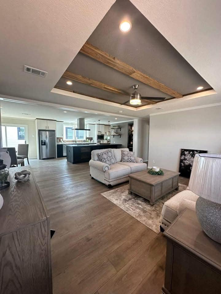 Contemporary open living area with light wood floors, cozy sofas, and a rustic coffee table. A modern kitchen is visible in the background. Warm, inviting ambiance.