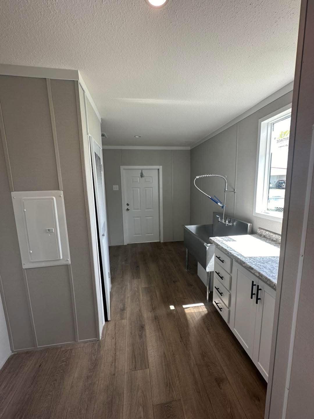 Narrow room with wood flooring, gray walls, and a door at the end. Right side has a large metal sink, white cabinets, and a window. Bright, minimalist feel.