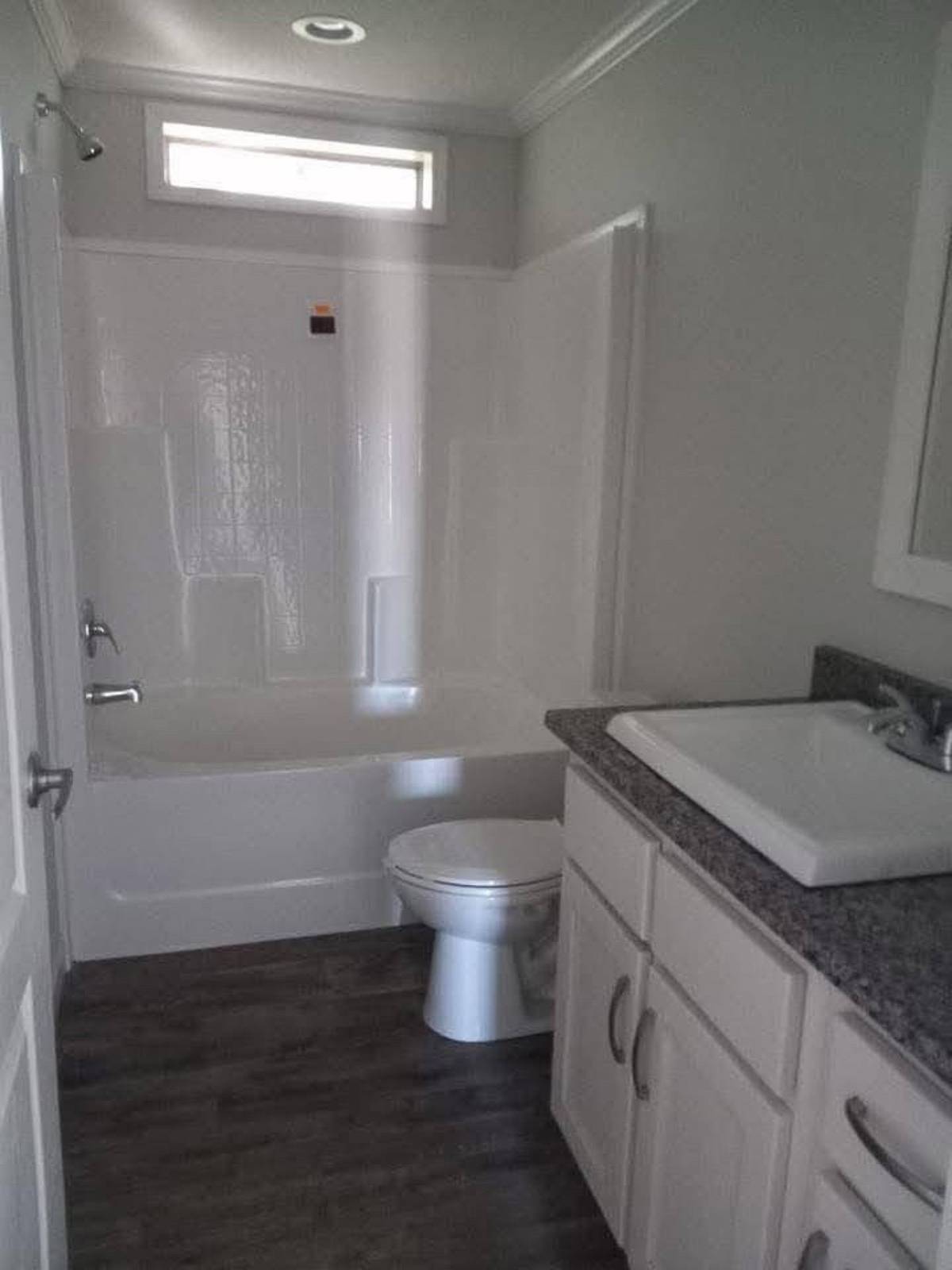 Narrow bathroom with wood floor, white bathtub-shower combo, small window, toilet, and gray granite countertop sink with white cabinets. Cozy and neat.