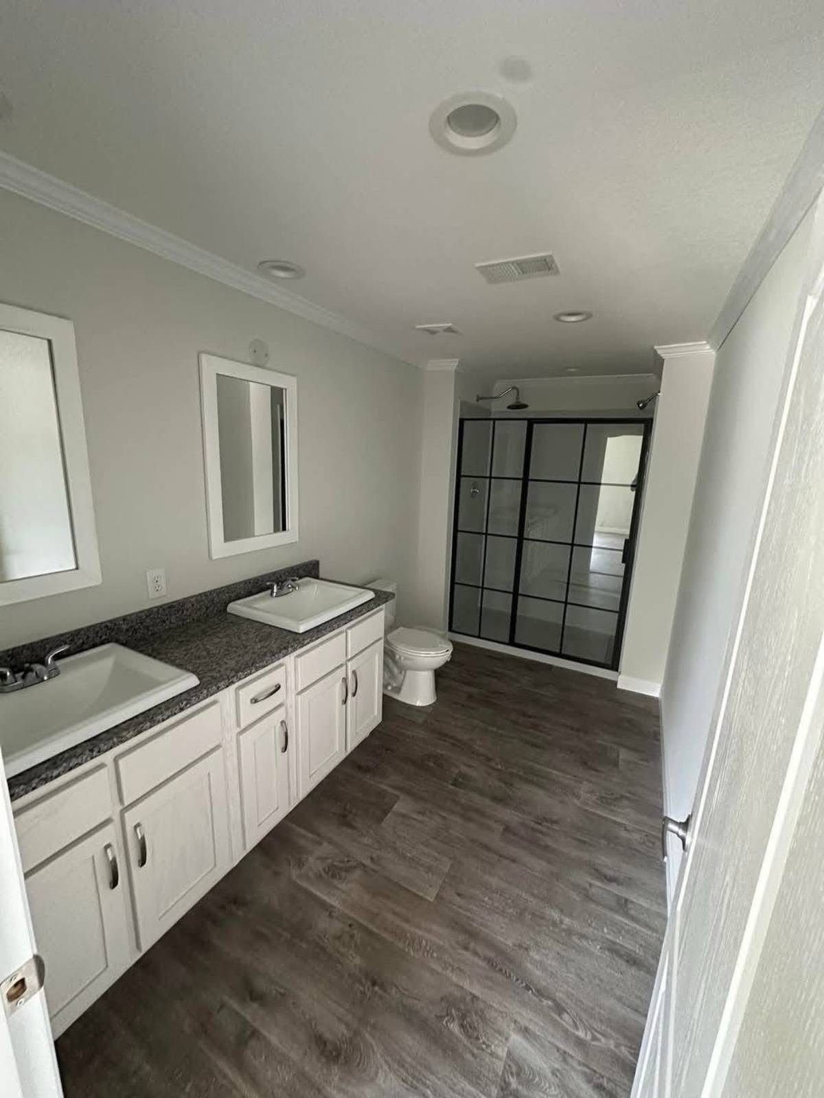 A modern bathroom with a double sink vanity on the left, granite countertop, white cabinets, a toilet in the center, and a shower with black framed glass doors. The floor is wood-patterned, creating a clean and fresh ambiance.