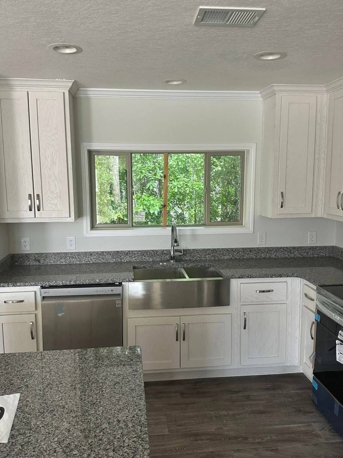 A modern kitchen with white cabinets, granite countertops, and a large stainless steel sink. A window above the sink offers a view of lush greenery.
