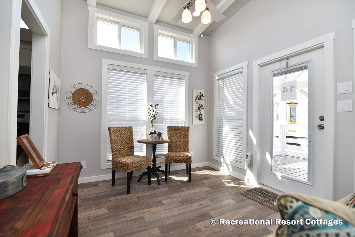 Bright cozy room with high ceilings, featuring two wicker chairs and a round table by large windows. Soft light creates a serene atmosphere.