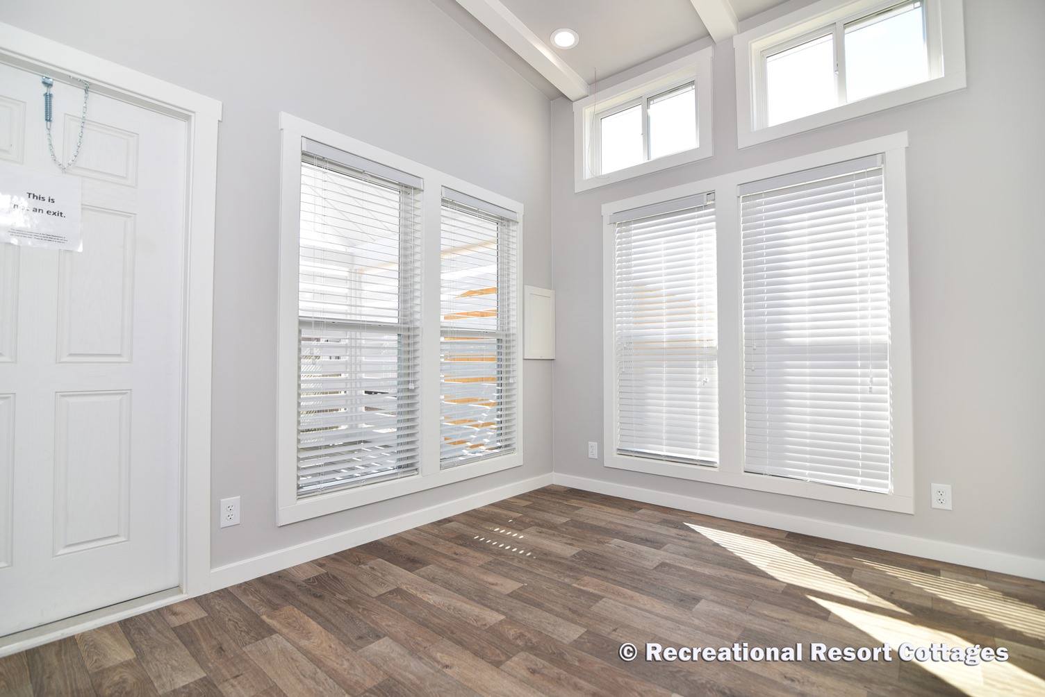 Bright room with two large sets of windows featuring white blinds, light gray walls, and wood-patterned flooring. The atmosphere is airy and modern.