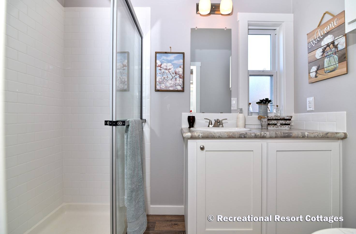 Modern bathroom with a glass-enclosed shower, white tile walls, and a gray towel. Vanity with marble countertop, mirror, and window, evoking a cozy feel.