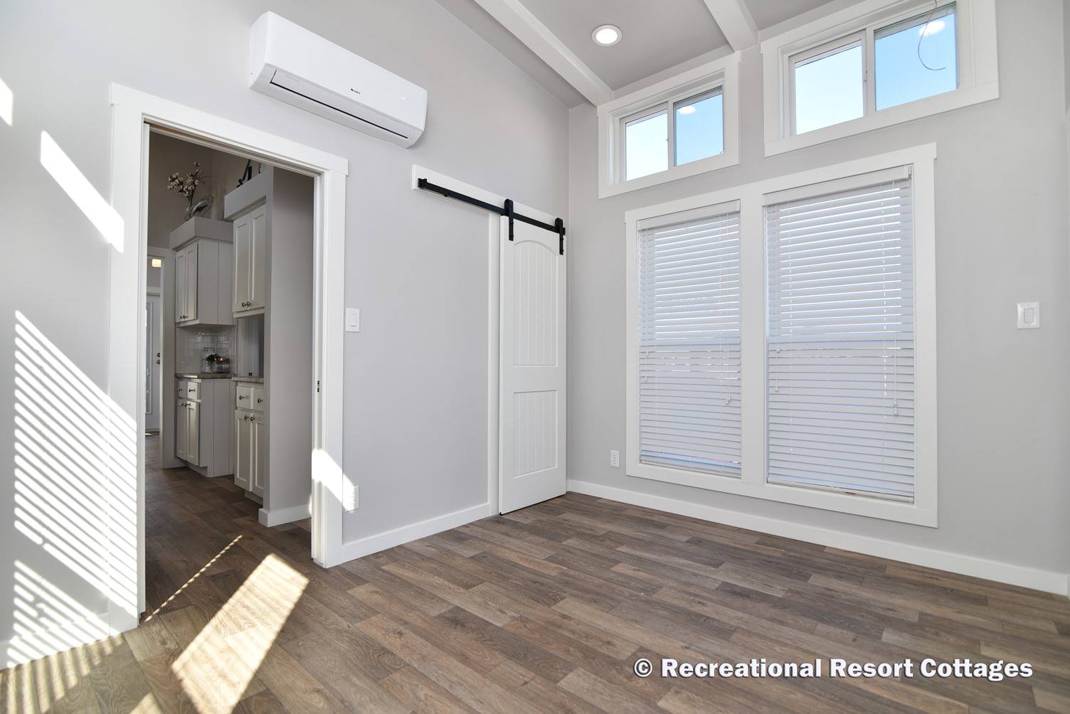 Spacious room with light gray walls and wood flooring, featuring large windows with blinds, a sliding barn door, and an adjacent kitchen entrance.
