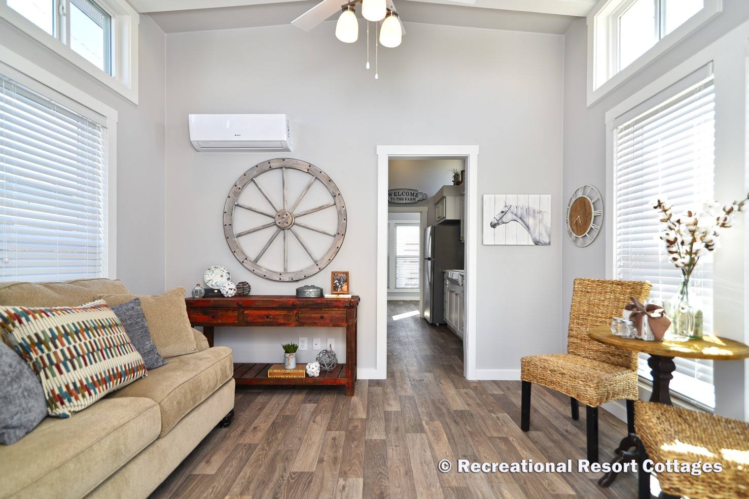 A cozy living room with neutral tones, featuring a beige sofa with colorful pillows, a rustic table with decor, and a large wheel wall art. Light floods in through large windows with blinds, creating a warm and inviting atmosphere. A small dining area with wicker chairs and a round table sits to the right, while an open doorway leads to a bright kitchen.