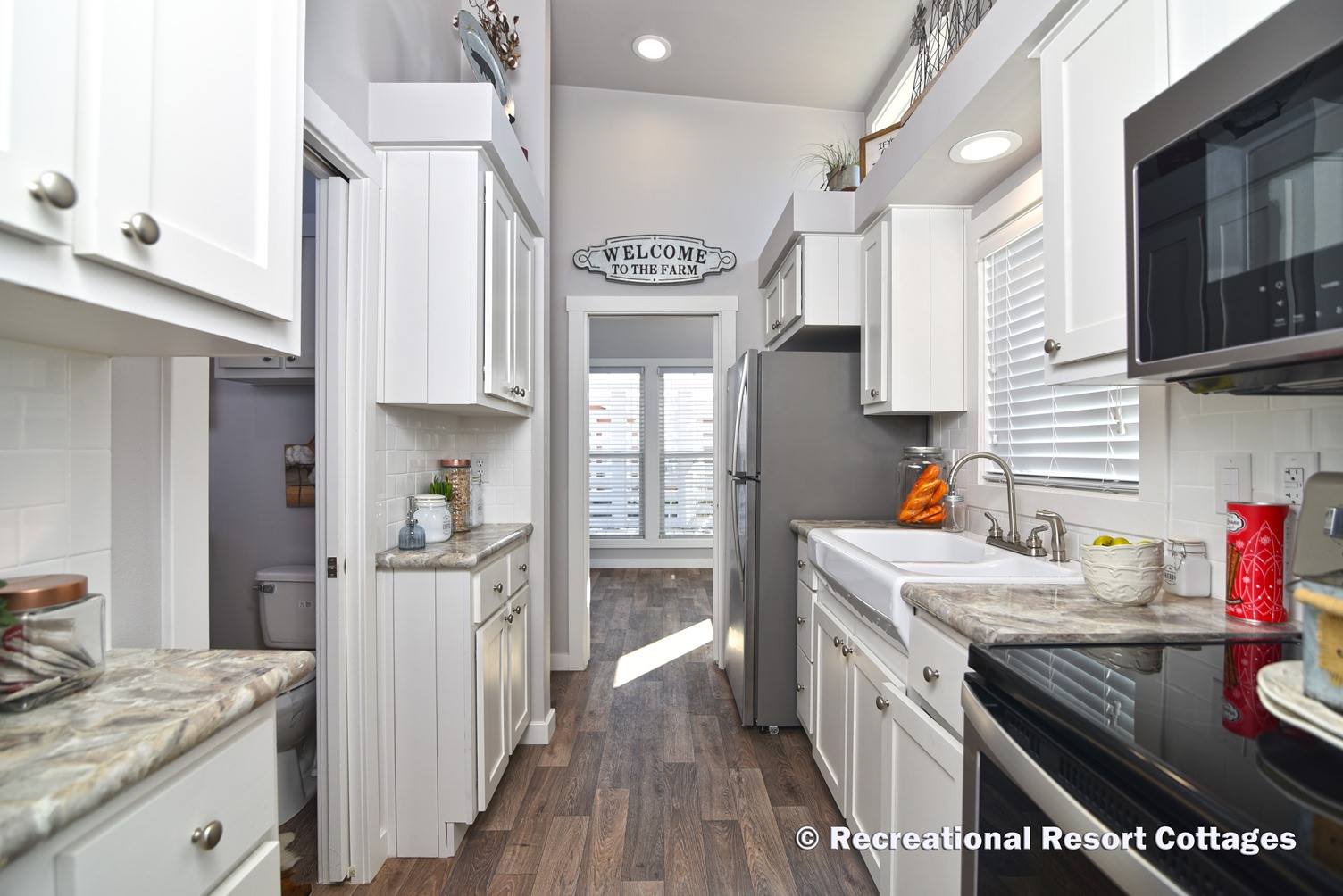 A cozy, narrow kitchen with white cabinetry, farmhouse sink, and granite countertops. Sunlight streams through a door with a "Welcome to the Farm" sign.