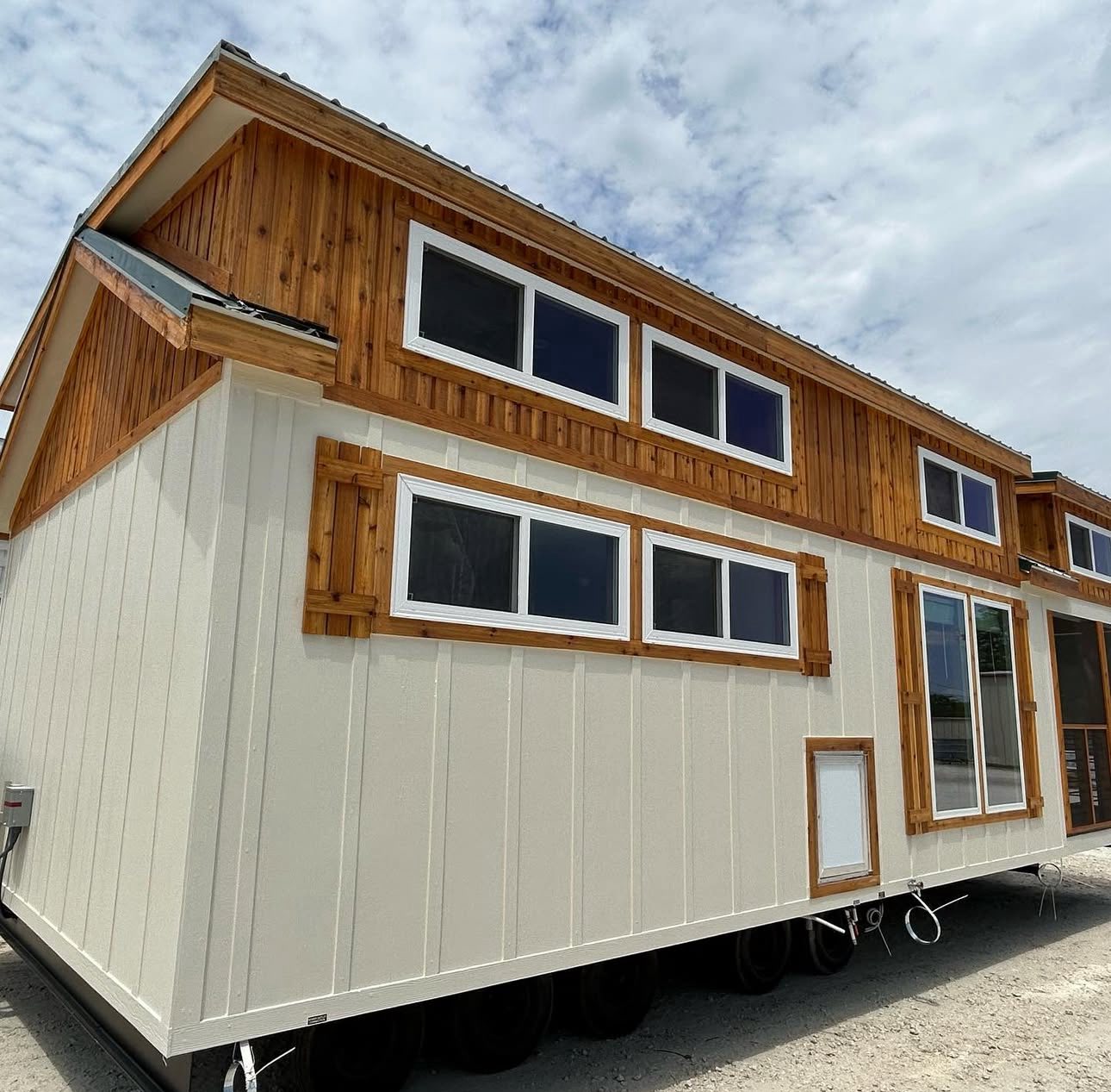 A beige tiny house with cedar paneling accents and multiple narrow windows sits on wheels under a cloudy sky, suggesting mobility and minimalism.