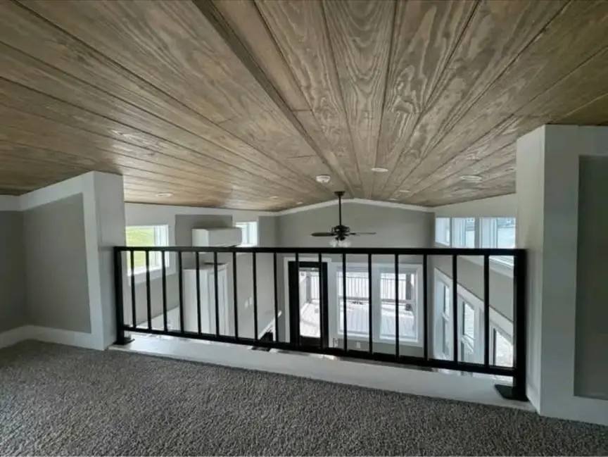 Modern loft space with a wooden ceiling, gray walls, and a black metal railing. Sunlight streams through large windows, creating an airy feel.