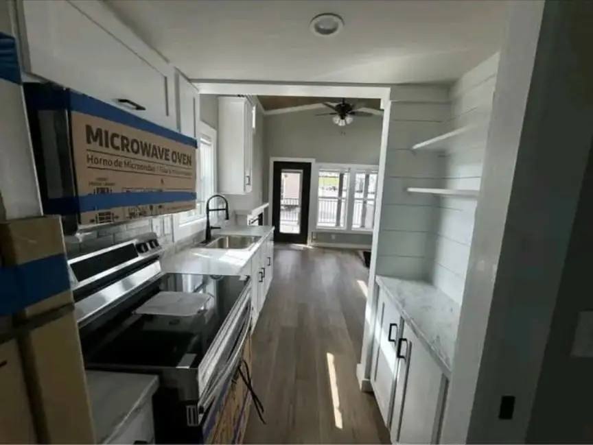 Modern kitchen with white cabinets, stainless appliances, and light wood flooring. Sunlit dining area with large windows is visible in the background.